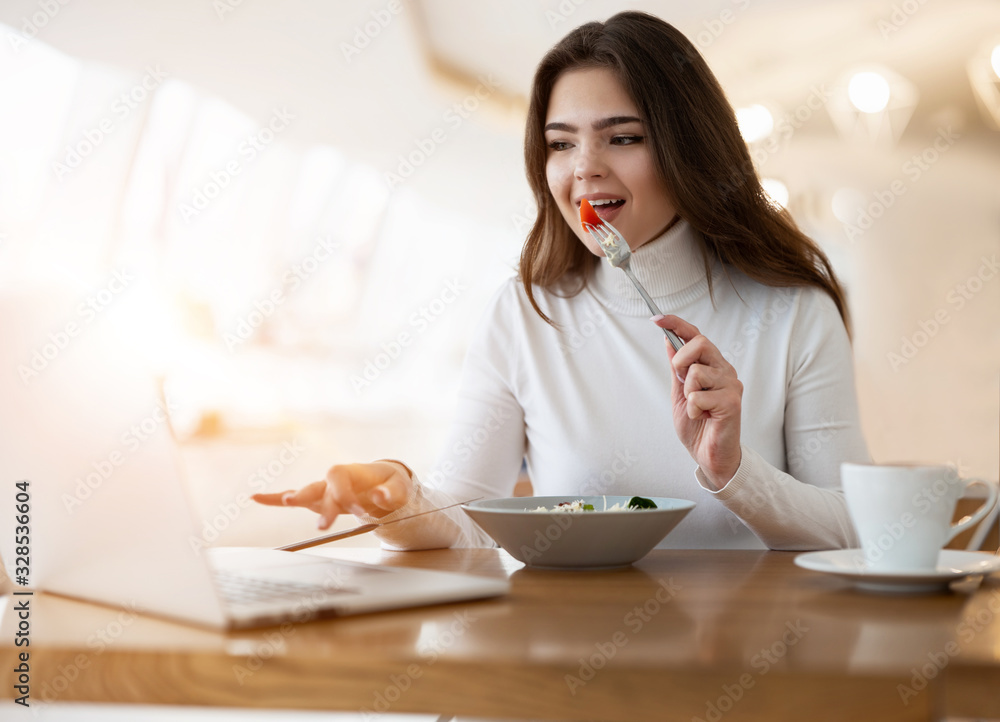 young positive beautiful woman works in her laptop biting tomatoe on the fork while eating salad in cafe during lunch time, multitasking concept young positive beautiful woman works in her laptop biting tomatoe on the fork while eating salad in cafe during lunch time, multitasking concept