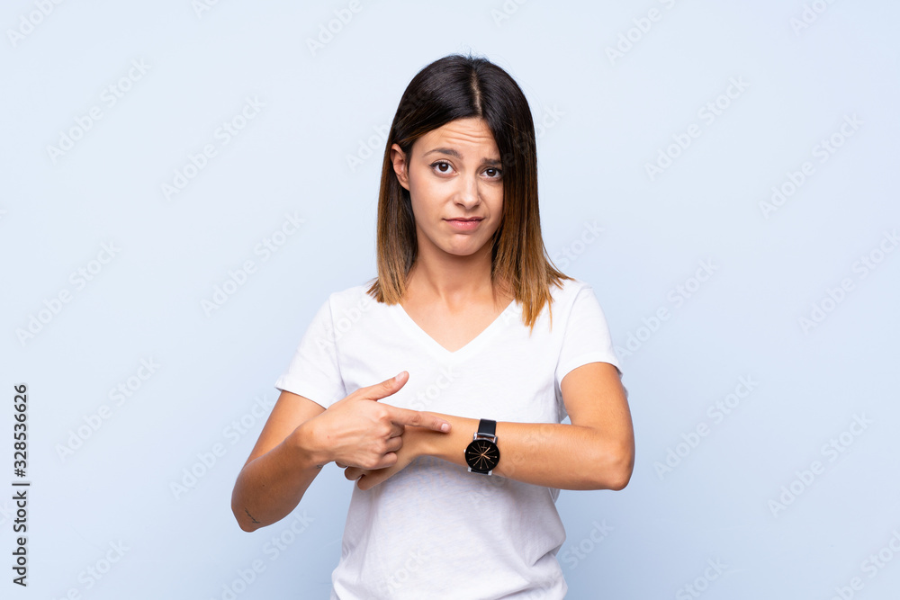 Young woman over isolated blue background showing the hand watch with serious expression Young woman over isolated blue background showing the hand watch with serious expression