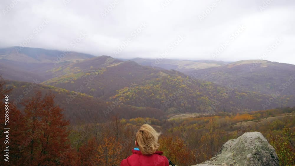 Female hiker in red taking a rest on a rock in the mountains. Windy cloudy day. Girl enjoying the view. Travel and healthy lifestyle outdoors in fall season. Vertical camera movement. Female hiker in red taking a rest on a rock in the mountains. Windy cloudy day. Girl enjoying the view. Travel and healthy lifestyle outdoors in fall season. Vertical camera movement.