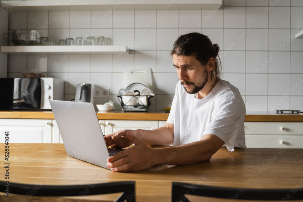 Freelancer working from home on the kitchen and using laptop. Bearded man working with a laptop and reading good news. Handsome successful self entrepreneur sitting and working at his modern home. Freelancer working from home on the kitchen and using laptop. Bearded man working with a laptop and reading good news. Handsome successful self entrepreneur sitting and working at his modern home.