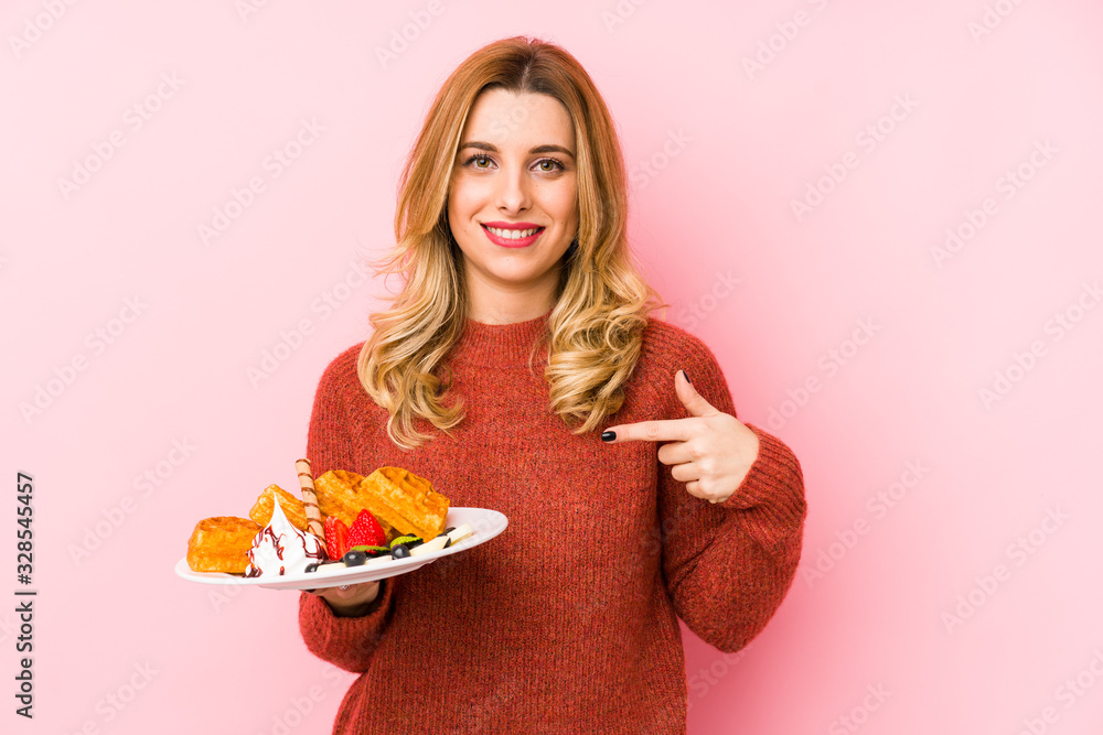 Young blonde woman eating a waffle dessert isolated person pointing by hand to a shirt copy space, proud and confident Young blonde woman eating a waffle dessert isolated person pointing by hand to a shirt copy space, proud and confident