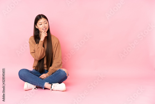 Young asian woman sitting on the floor isolated on pink background looking to the side © luismolinero