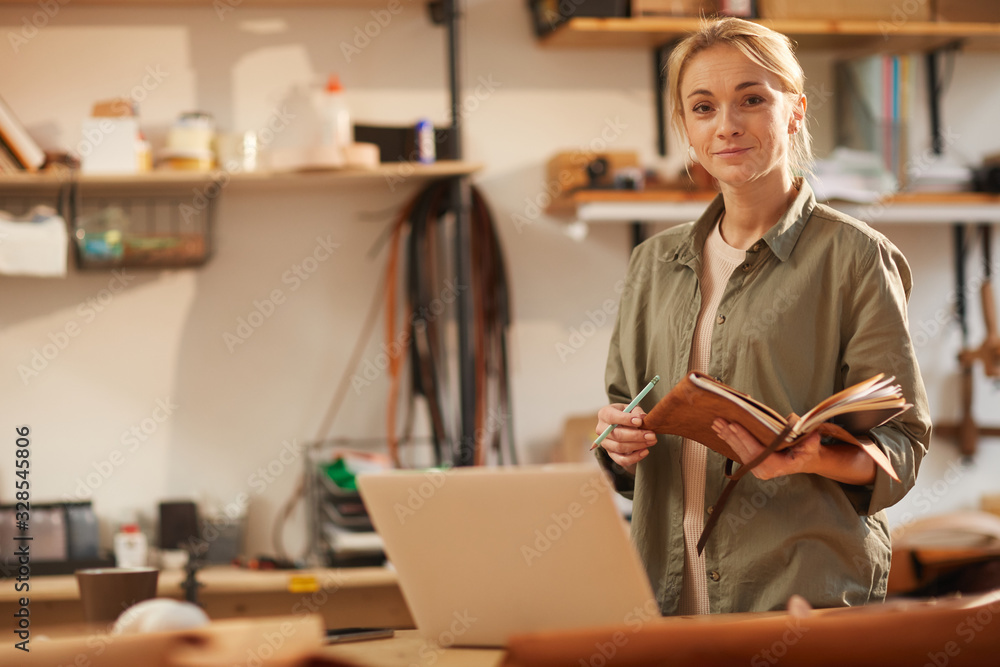 Horizontal portrait of attractive young woman wearing casual outfit standing at table with laptop on it holding notebooks looking at camera Horizontal portrait of attractive young woman wearing casual outfit standing at table with laptop on it holding notebooks looking at camera
