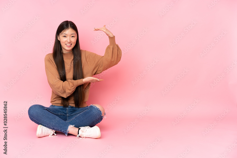 Young asian woman sitting on the floor isolated on pink background holding copyspace to insert an ad Young asian woman sitting on the floor isolated on pink background holding copyspace to insert an ad