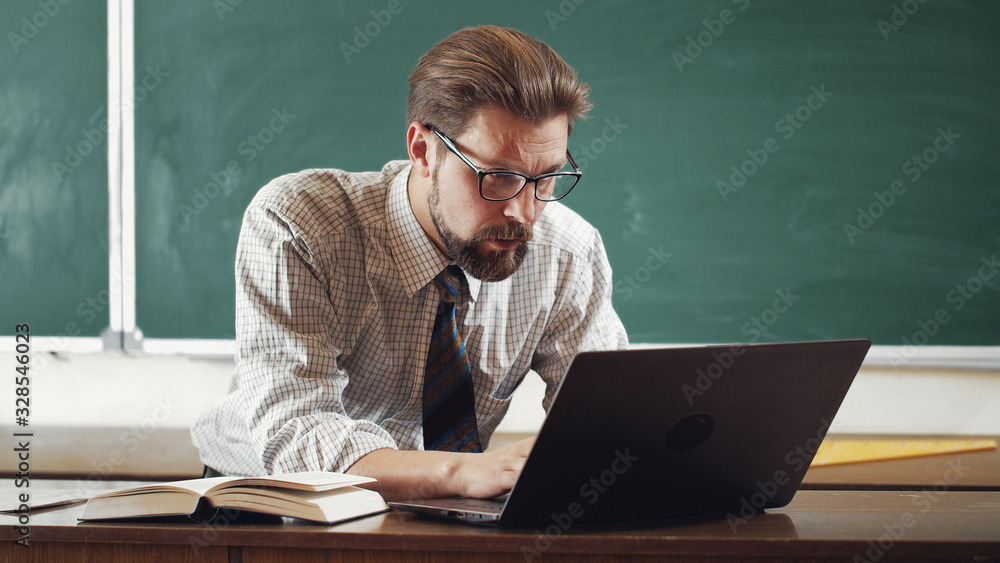 Serious mature bearded teacher wearing shirt, tie, and spectacles working on laptop in classroom Serious mature bearded teacher wearing shirt, tie, and spectacles working on laptop in classroom