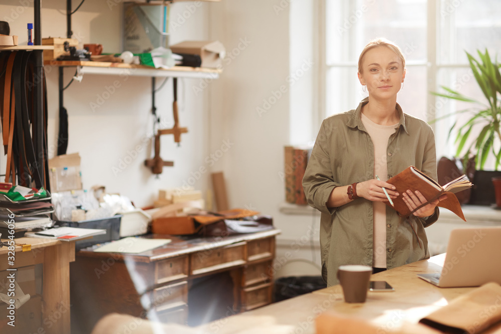 Horizontal portrait of young Caucasian woman wearing casual outfit standing at table with laptop on it holding notebooks looking at camera Horizontal portrait of young Caucasian woman wearing casual outfit standing at table with laptop on it holding notebooks looking at camera