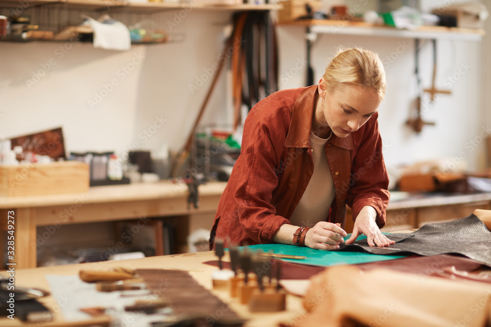 Horizontal portrait of young Caucasian craftswoman creating something using piece of leather Horizontal portrait of young Caucasian craftswoman creating something using piece of leather