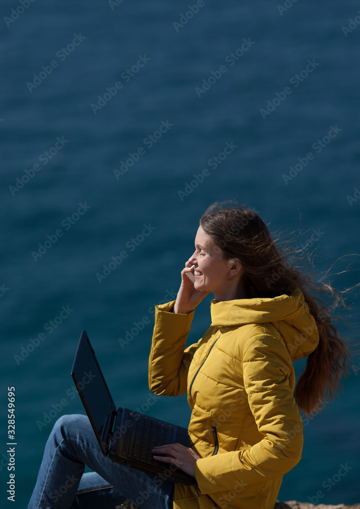 Technology and travel. Work in the open air. The concept of freelancing. A pretty young woman in a yellow jacket uses a laptop , talks on the phone and smiles with a view of the sea and the sky.