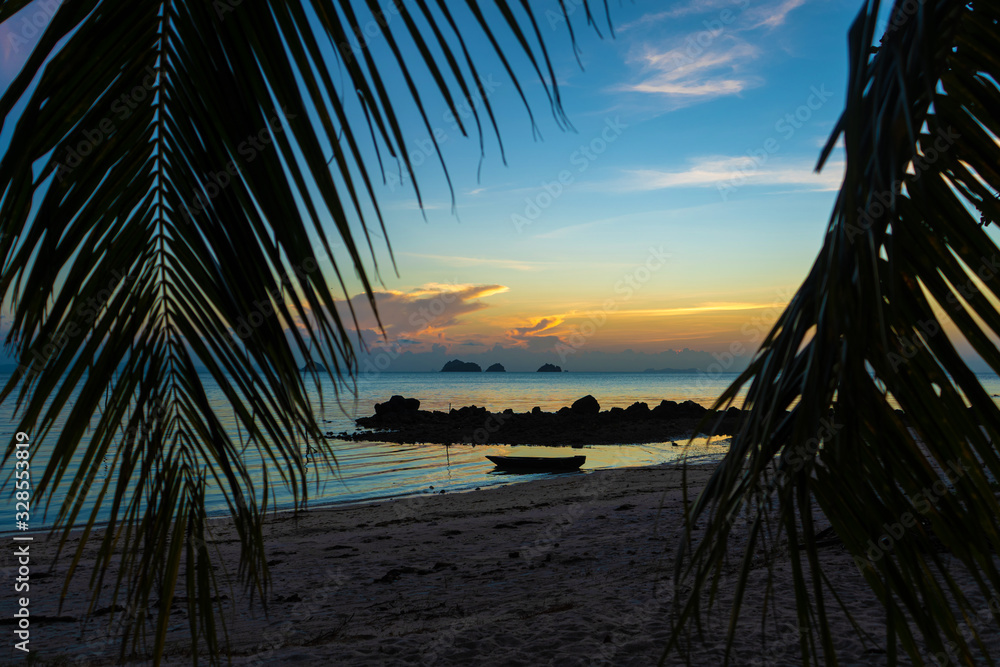 View through the leaves of palm trees on the ocean. There is a wooden boat on the water. Sunset. Sand beach. Romance View through the leaves of palm trees on the ocean. There is a wooden boat on the water. Sunset. Sand beach. Romance