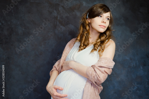 Pregnant young woman touching her belly. Beautiful expectant mom on dark background. Pregnancy, maternity clothes concept.