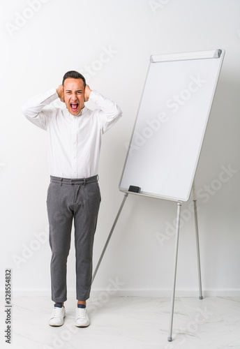 Young latin coaching man with a whiteboard isolated covering ears with hands trying not to hear too loud sound.