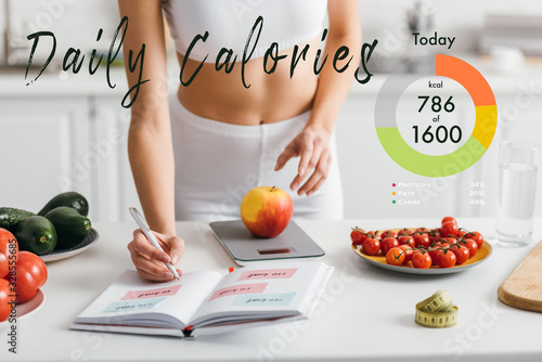 Cropped view of fit sportswoman writing calories while weighing apple on kitchen table, daily calories illustration