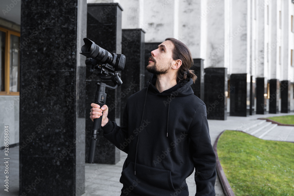 Young Professional videographer holding professional camera on 3-axis gimbal stabilizer. Pro equipment helps to make high quality video without shaking. Cameraman wearing black hoodie making a videos. Young Professional videographer holding professional camera on 3-axis gimbal stabilizer. Pro equipment helps to make high quality video without shaking. Cameraman wearing black hoodie making a videos.