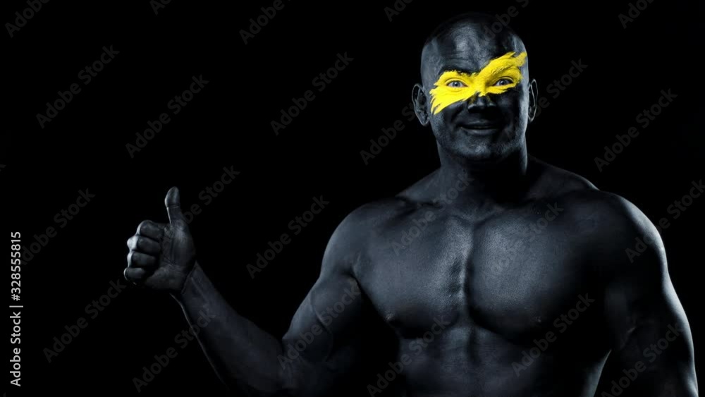 Man sports fan and bodybuilder athlete with yellow color on face art and black body paint. Colorful portrait of the guy with bodyart. Guy showing thumbs up sign with fingers. Man sports fan and bodybuilder athlete with yellow color on face art and black body paint. Colorful portrait of the guy with bodyart. Guy showing thumbs up sign with fingers.