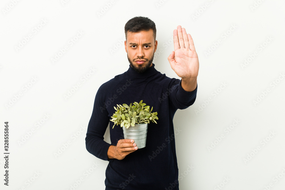 Young south-asian man holding a plant standing with outstretched hand showing stop sign, preventing you. Young south-asian man holding a plant standing with outstretched hand showing stop sign, preventing you.