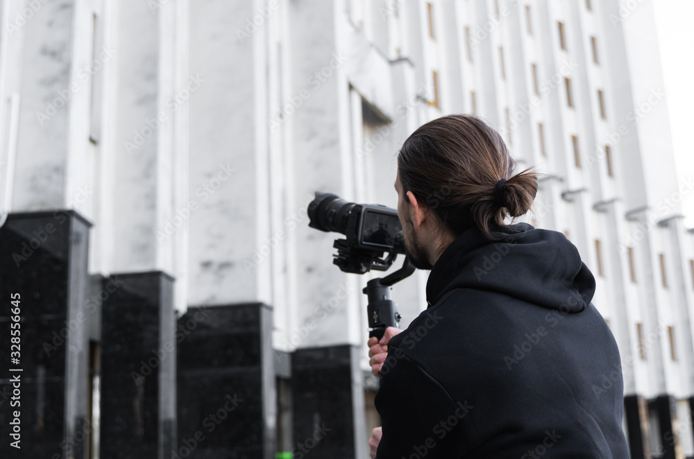 Young Professional videographer holding professional camera on 3-axis gimbal stabilizer. Pro equipment helps to make high quality video without shaking. Cameraman wearing black hoodie making a videos. Young Professional videographer holding professional camera on 3-axis gimbal stabilizer. Pro equipment helps to make high quality video without shaking. Cameraman wearing black hoodie making a videos.