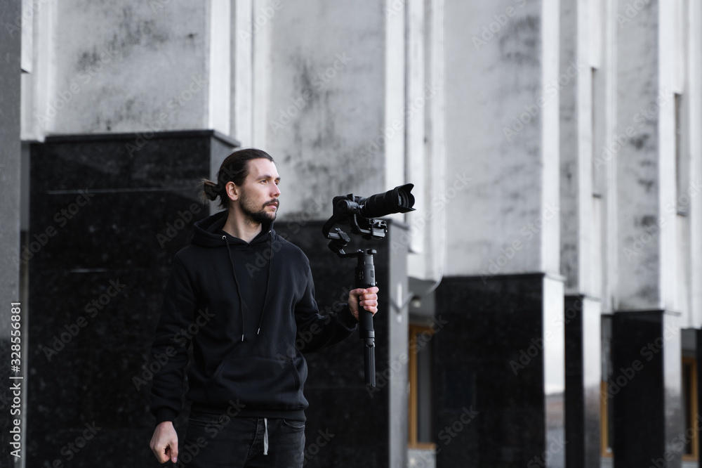 Young Professional videographer holding professional camera on 3-axis gimbal stabilizer. Pro equipment helps to make high quality video without shaking. Cameraman wearing black hoodie making a videos. Young Professional videographer holding professional camera on 3-axis gimbal stabilizer. Pro equipment helps to make high quality video without shaking. Cameraman wearing black hoodie making a videos.