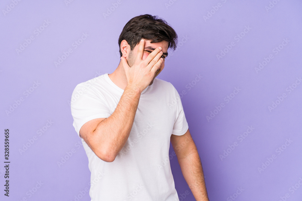 Young man isolated on purple background blink at the camera through fingers, embarrassed covering face. Young man isolated on purple background blink at the camera through fingers, embarrassed covering face.
