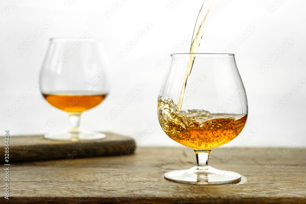 Cognac \ whiskey in cognac glass on wooden table, golden color spirits splashing on white background Cognac \ whiskey in cognac glass on wooden table, golden color spirits splashing on white background