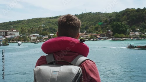 Tourist man stands on deck looking at the island's harbour while long tail boats pass by