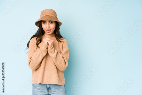 Young indian woman wearing a hat isolated on blue background praying for luck, amazed and opening mouth looking to front.