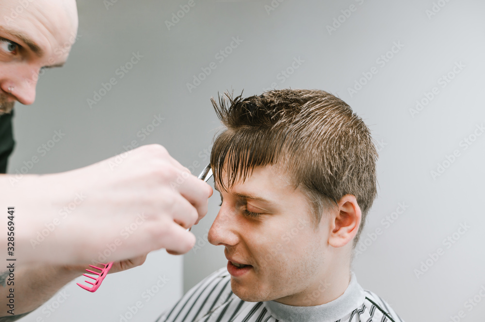 Obraz premium Closeup photo of a young blond man clipping at a men's bar in a barber's favorite. Professional haircutter cuts client's hair scissors with blond hair. Men's haircut concept.