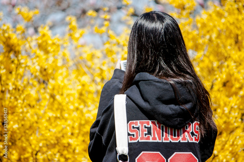 Back of Young Woman With Long Black Hair With Senior Jacket Looking at her Phone Against Yellow Bush photo