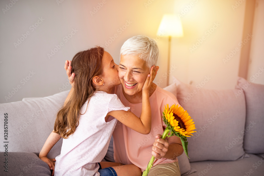 Cute girl giving a bunch of flowers to her grandmother sitting on the couch. Happy grandmother hugging small cute grandchild thanking for flowers presented. Darling granddaughter always remembers - obrazy, fototapety, plakaty 