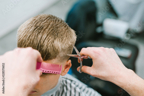Professional hairdresser cuts the hair of a young blond man with scissors and a comb. Closeup photo of a barber's hands in the process of scissors. Creating male hairstyle background.
