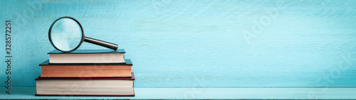 A magnifying glass on a stack of books on a blue wooden bookshelf with copy space web banner photo