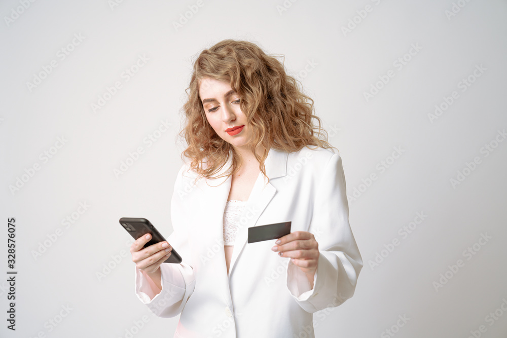 Confident young curly woman in white blazer holds credit card in one hand and phone in other, isolated over grey background Confident young curly woman in white blazer holds credit card in one hand and phone in other, isolated over grey background