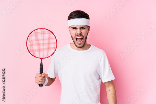 Young caucasian man playing badminton isolated screaming very angry and aggressive. © Asier