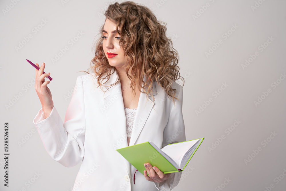 Beautiful curly woman top manager dressed in elegant white suit holds opened notebook in one hand and pink pen in other, looking at the left side Beautiful curly woman top manager dressed in elegant white suit holds opened notebook in one hand and pink pen in other, looking at the left side