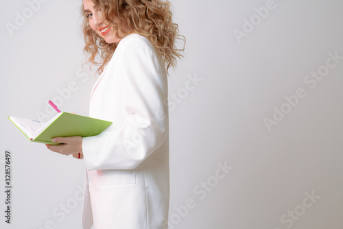 Happy curly woman teacher in white blazer holds notebook and pen in her hands and looking at the camera over her shoulder, isolated over grey background