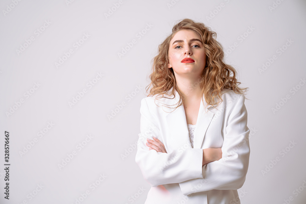 Waist up portrait of a curly blonde girl in white blazer who stands with crossed hands and looking seriously at the camera isolated over grey background