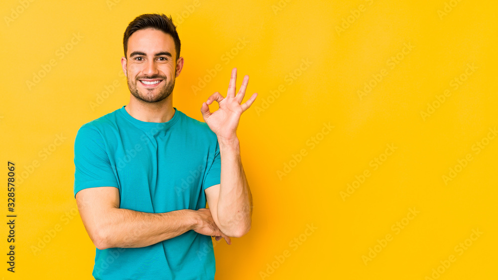 Young caucasian man isolated on yellow bakground winks an eye and holds an okay gesture with hand. Young caucasian man isolated on yellow bakground winks an eye and holds an okay gesture with hand.