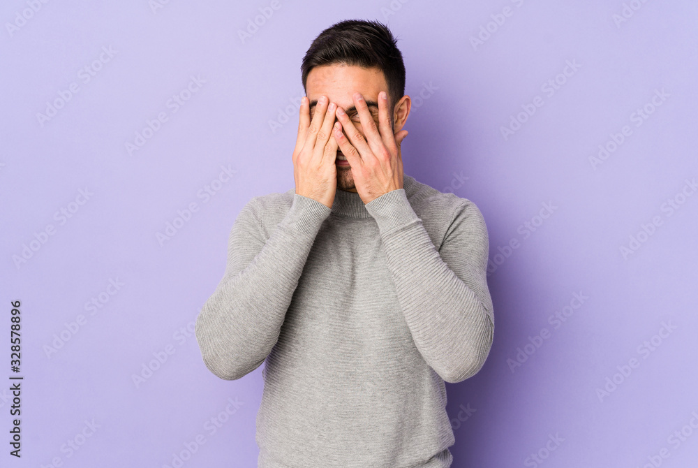 Young caucasian man isolated on purple background blink at the camera through fingers, embarrassed covering face. Young caucasian man isolated on purple background blink at the camera through fingers, embarrassed covering face.