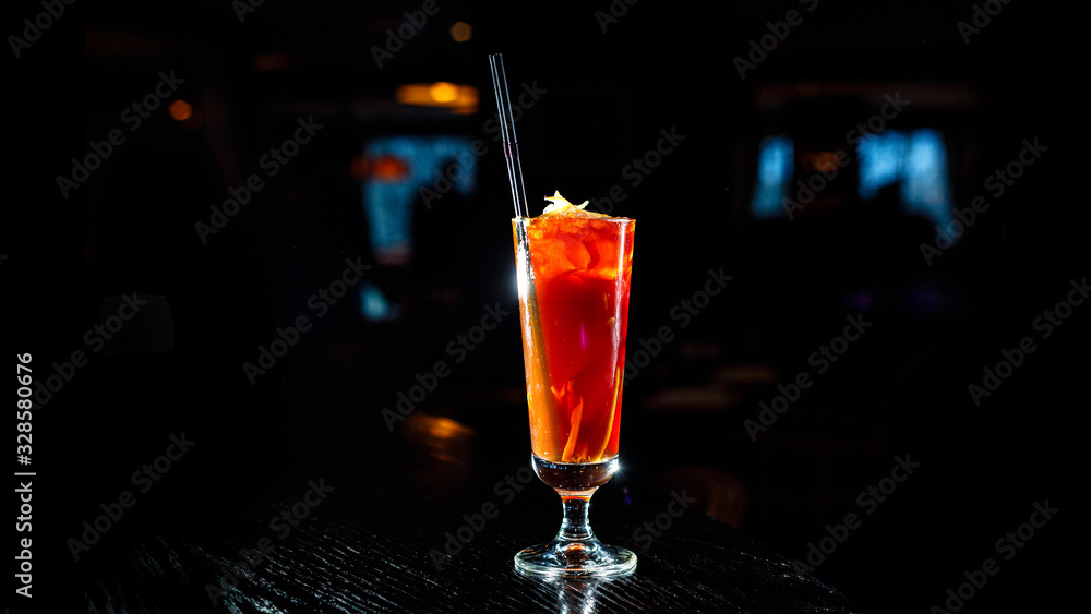 A red cocktail in a tall glass stands on the counter of a dark bar. A red cocktail in a tall glass stands on the counter of a dark bar.