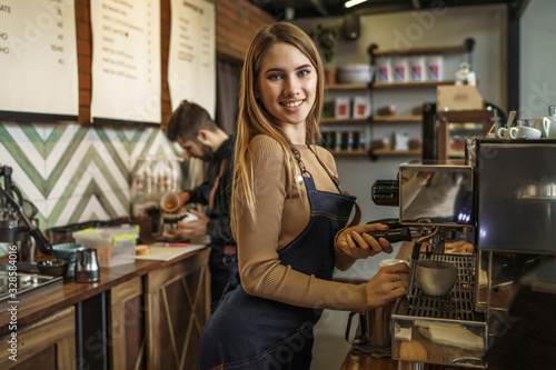 Professional female barista holding metal jug warming milk using the coffee machine. Positive smiling woman preparing coffee at counter.