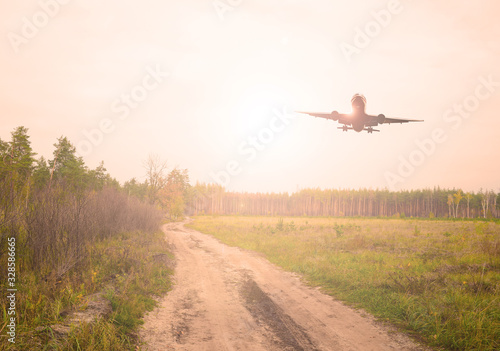 Plane over a sand road in a green field near the forest. The sun is shining
