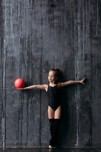 Girl gymnast posing with a red ball. Gymnast dressed in sportswear with golf. On the background is a black textured wall.