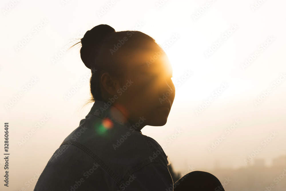 Silhouette of the thinking Asian woman sitting on a roof with city background at sunrise. 