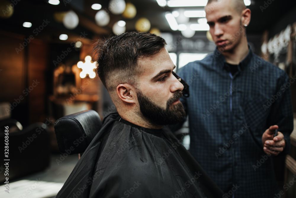 Good looking young adult man getting a hair and beard styling and dressing treatment by professional hairstylist. Good looking young adult man getting a hair and beard styling and dressing treatment by professional hairstylist.