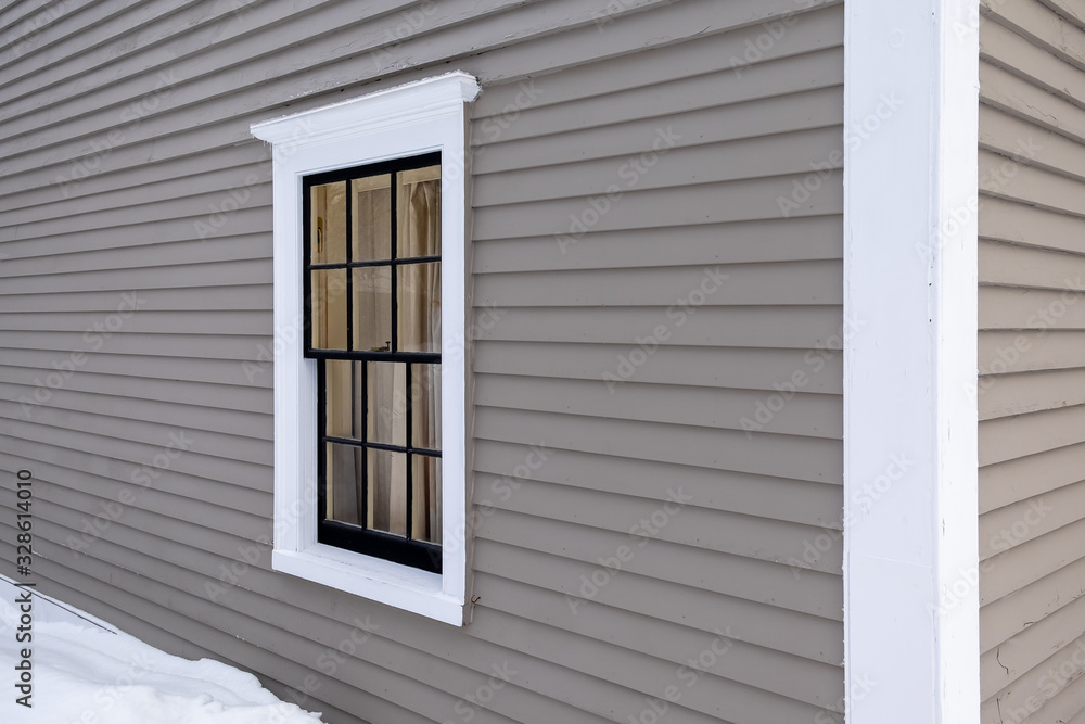 Fototapeta premium The exterior wall of a grey colored building made of narrow wooden clapboard with a large drift of snow. There's a double hung closed window in the wall with white trim and black glass dividers.