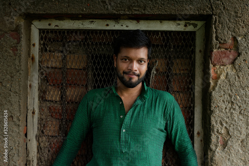 Portrait of an young and handsome Indian Bengali brunette man standing in front of a vintage house window wearing green Indian traditional ethnic cloths. Indian lifestyle and fashion © abir