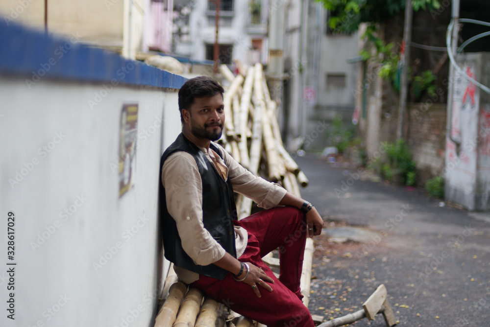 Portrait of an young and handsome Indian Bengali brunette beard man sitting on a pile of bamboos on roadside wearing black jacket and  white shirt. Indian lifestyle and fashion