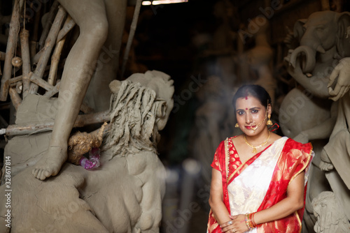 Portrait of an young and beautiful Indian Bengali brunette woman in red and white traditional ethnic sari in front of the clay idol of Hindu Bengali goddess Durga. Indian culture, religion and fashion photo