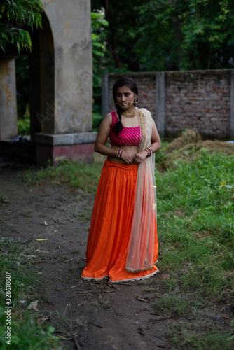 Young and beautiful Indian Bengali brunette woman standing thoughtfully in green nature in front of a vintage house wearing Indian traditional ethnic vibrant skirt blouse. Indian lifestyle and fashion