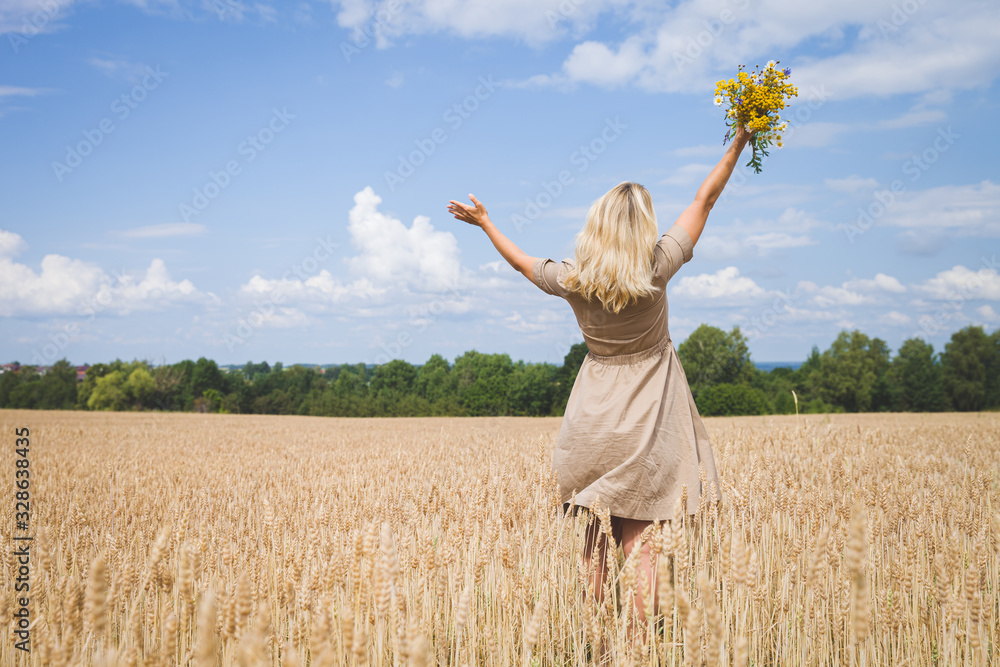 Happy girl with a bouquet of wild flowers walking in a wheat field. Girl with flowers and a wheat field on a background of blue sky.View from the back of a beautiful girl with hands up. Summer concept Happy girl with a bouquet of wild flowers walking in a wheat field. Girl with flowers and a wheat field on a background of blue sky.View from the back of a beautiful girl with hands up. Summer concept