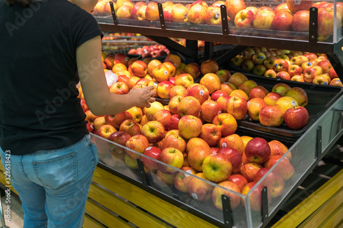 Fresh healthy fruits on shelves in supermarket, with female buyer choosing red apples